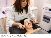 Female cosmetician in white uniform using applying cleansing cream, with brush on face of woman covered with towel and lying face up eyes closed in beauty center during makeup session. Beauty specialist applying cosmetics on female client in beauty salon. Стоковое фото, агентство Ingram Publishing / Фотобанк Лори