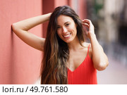 A joyful woman with long hair smiles brightly, playfully posing with one hand in her hair. She wears a vibrant red dress, complementing the charming urban backdrop, creating a lively scene. A smiling woman with long hair in a vibrant red dress posing gracefully outdoors. Стоковое фото, агентство Ingram Publishing / Фотобанк Лори