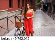 A fashionable woman in a vibrant red dress strolls with her ebike along a charming cobblestone street, surrounded by elegant architecture, enjoying the relaxed atmosphere of the city. A Stylish Woman Dressed in a Red Dress Walking Gracefully with a ebike through an Urban Setting. Стоковое фото, агентство Ingram Publishing / Фотобанк Лори