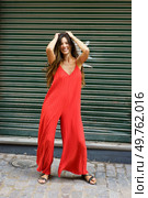 A vibrant and energetic young woman dressed in a stylish red jumpsuit confidently poses against a striking green shutter backdrop, embodying a sense of fun and spontaneity in her expression. A Stylish and Trendy Young Woman in a Bright Red Jumpsuit Posing Happily Outdoors. Стоковое фото, агентство Ingram Publishing / Фотобанк Лори