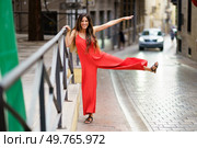 A chic and stylish woman in an eyecatching vibrant red dress, strikes a playful pose atop a railing in the midst of a charming and picturesque city street filled with life and color. An Elegant Woman Dressed in a Stunning Red Dress Posing Gracefully on a Street Railing. Стоковое фото, агентство Ingram Publishing / Фотобанк Лори