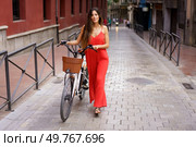 A young woman dressed in a vibrant red outfit stands confidently with her stylish ebike parked nearby on a lively urban street, showcasing her natural elegance, fashionable flair, and unique style. Charming Young Woman Riding an ebike in a Lively Urban Street Setting Full of Life. Стоковое фото, агентство Ingram Publishing / Фотобанк Лори