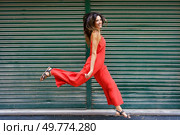 A vibrant and energetic woman, dressed stylishly in an eyecatching red dress, happily showcases her playful spirit while cheerfully walking against a lush green backdrop, full of life and color. An Energetic Woman Dressed in a Striking Red Dress is Joyfully Walking Outdoors in the City. Стоковое фото, агентство Ingram Publishing / Фотобанк Лори