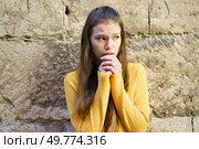 A young woman, dressed in a cozy yellow sweater, stands against an old, rustic stone wall, appearing worried. Her expression poignantly captures a moment filled with anxiety or deep contemplation. A Young Woman Dressed in a Yellow Sweater is Expressing Concern Against a Textured Wall. Стоковое фото, агентство Ingram Publishing / Фотобанк Лори