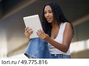 A vibrant young woman with curly hair beams with joy as she interacts with her tablet, embodying happiness while enjoying a casual day in a lively outdoor setting, surrounded by nature and life. A Young Woman is Actively Engaged with Her Tablet While in a Casual, Relaxed Setting. Стоковое фото, агентство Ingram Publishing / Фотобанк Лори