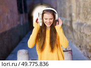 In a vibrant urban alley with artistic murals, a cheerful young woman in a bright yellow sweater smiles joyfully, immersed in the rhythm of her favorite music while wearing headphones. A Joyful Young Woman Enjoying Listening to Music in a Colorful Urban Setting Outdoors. Стоковое фото, агентство Ingram Publishing / Фотобанк Лори