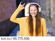 A cheerful and vibrant young woman, dressed in a bright yellow sweater and wearing headphones, joyfully expressing her passion for music amidst the lively atmosphere of an urban environment. A Joyful Young Woman is Truly Enjoying Music in a Dynamic Urban Setting with Delight. Стоковое фото, агентство Ingram Publishing / Фотобанк Лори