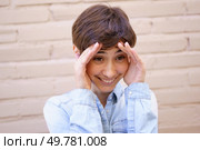 A cheerful individual with short hair shows surprise and joy, playfully placing their hands on their head against a warm beige brick background that enhances the mood of happiness. A Joyful Young Person Expressing Genuine Surprise and Delight in a Vibrant Setting. Стоковое фото, агентство Ingram Publishing / Фотобанк Лори