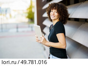A joyful woman with curly hair stands outdoors, holding a tablet in her hands and smiling radiantly, showcasing a modern lifestyle that reflects a sense of happiness and connection with nature. A cheerful woman with curly hair joyfully holding a tablet while enjoying the outdoors. Стоковое фото, агентство Ingram Publishing / Фотобанк Лори