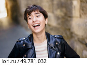 A lively and cheerful young woman with trendy short hair, wearing a stylish leather jacket, is laughing joyfully as she poses against a backdrop of an artistic stone wall in an urban environment. A Joyful Young Woman Laughing Cheerfully in a Lively Urban Setting with Vibrant Colors. Стоковое фото, агентство Ingram Publishing / Фотобанк Лори