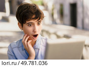 A young woman with a stylish short haircut looks at her laptop outdoors, her face showing surprise and curiosity about an unexpected online discovery, reflecting her engagement with the digital world. A Young Woman Looks Surprised and Reacts While Viewing Her Laptop Screen Outdoors. Стоковое фото, агентство Ingram Publishing / Фотобанк Лори