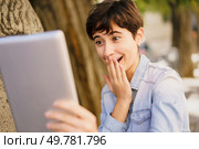 A young woman with a stylish short haircut displays a mixture of surprise and genuine joy as she gazes at her tablet, fully capturing authentic emotions while enjoying a relaxed outdoor setting. A Young Woman is Expressing Surprise and Delight in Reaction to Her Tablet Screen Display. Стоковое фото, агентство Ingram Publishing / Фотобанк Лори