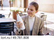 A smiling woman sitting at a caf table, admiring her stylish white water bottle in a vibrant outdoor setting. A Woman Happily Enjoying Her Colorful Water Bottle While Seated in a Cafeteria Setting. Стоковое фото, агентство Ingram Publishing / Фотобанк Лори