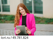 A cheerful young woman in a pink blazer is enjoying her outdoor leisure time. She holds a tablet while appreciating the fresh air, bright sunlight, and beautiful greenery around her. A Cheerful Young Woman Happily Engaged with Her Tablet in a Beautiful Outdoor Setting. Стоковое фото, агентство Ingram Publishing / Фотобанк Лори