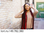 A stylish young woman with long, beautifully braided hair listens to music through her headphones, completely immersed in her own world, set against a vibrant urban background full of life. A young woman joyfully listens to music through headphones in a vibrant urban setting. Стоковое фото, агентство Ingram Publishing / Фотобанк Лори