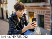 A youthful individual sporting a fashionable short haircut, and a stylish leather jacket is expressing a sense of astonishment as they gaze intently at their smartphone while enjoying the outdoors. A Young Person Reacting Emotionally to Surprising Content on Their Mobile Device. Стоковое фото, агентство Ingram Publishing / Фотобанк Лори