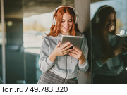 A cheerful young woman with vibrant red curly hair is joyfully enjoying her music streaming on a sleek tablet while wearing stylish headphones, all while she stands in a lively urban setting. A young woman enjoys music on her tablet, wearing headphones and fully immersed in it. Стоковое фото, агентство Ingram Publishing / Фотобанк Лори