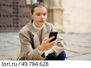 A stylish young woman sits outdoors, absorbed in her smartphone, immersed in digital interaction. Textured walls in the background enhance the modern life essence and connectivity. A Young Woman is Engaged with Her Smartphone While Sitting Outdoors in a Public Space. Стоковое фото, агентство Ingram Publishing / Фотобанк Лори