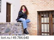 A fashionable and stylish young woman is sitting comfortably on a stone wall, completely focused on her smartphone, immersed in a charming urban environment characterized by beautiful brick walls. A young woman is using her smartphone in an urban outdoor setting, amidst contemporary life. Стоковое фото, агентство Ingram Publishing / Фотобанк Лори