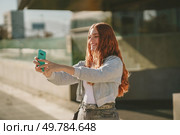 A cheerful young woman with long, flowing curly hair joyfully takes a selfie outdoors in the bright sunlight, framed by a lively urban backdrop full of dynamic energy and colors all around her. A Young Woman Enjoying Taking a Selfie While Outdoors in a Vibrant Urban Setting. Стоковое фото, агентство Ingram Publishing / Фотобанк Лори