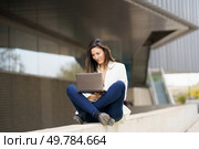 A young woman is comfortably seated on a ledge, deeply focused on her laptop in a vibrant contemporary urban environment, vividly showcasing the modern remote work lifestyle alongside technology. A Young Woman Concentrating While Working on a Laptop Outdoors in a Modern Urban Environment. Стоковое фото, агентство Ingram Publishing / Фотобанк Лори