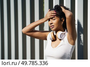 A young woman stands outdoors, wearing headphones, while expressing her emotional struggles against a backdrop that is distinctly modern and urban, reflecting contemporary life and its challenges. A woman wearing a white tank top and headphones thoughtfully faces challenges in the outdoors. Стоковое фото, агентство Ingram Publishing / Фотобанк Лори