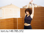 On a rooftop, a young woman with curly hair stretches her arms skyward, immersed in her yoga practice, embodying health and wellness in a vibrant urban setting full of energy and life. A Young Woman Engaged in Practicing Yoga Outdoors While Wearing Stylish Activewear. Стоковое фото, агентство Ingram Publishing / Фотобанк Лори