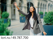 A stylish young woman with beautiful curly hair waves cheerfully while walking confidently through a vibrant urban area, showcasing her impeccable fashion sense and lively personality with every step. A Confident Young Woman Waving While Standing Outdoors in a Vibrant City Setting. Стоковое фото, агентство Ingram Publishing / Фотобанк Лори