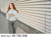 A cheerful young woman with curly red hair is outdoors, checking her smartphone in stylish casual wear. Modern architecture enhances the vibrant atmosphere of the lively urban environment around her. A young woman confidently holding her smartphone while standing against a modern wall. Стоковое фото, агентство Ingram Publishing / Фотобанк Лори