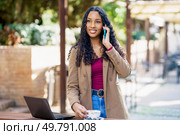 In a serene outdoor environment, a young woman is engaged in a focused phone call while standing next to her laptop, exuding confidence and determination as she multitasks effortlessly. A young woman sits outdoors, using her smartphone and laptop in a workspace setting. Стоковое фото, агентство Ingram Publishing / Фотобанк Лори