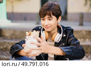A young woman in a trendy jacket sits on steps in a lively outdoor area, enjoying music through her headphones and a delicious dessert, creating a joyful vibe in a bustling urban setting. A Young Woman Fully Enjoying a Delicious Dessert While Wearing Headphones in an Urban Setting. Стоковое фото, агентство Ingram Publishing / Фотобанк Лори