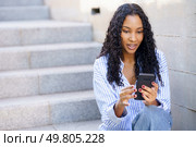 In a vibrant urban setting, a young woman with bouncy curly hair sits on outdoor steps, fully engaged with her smartphone, symbolizing the modern lifestyle many aspire to in their busy lives. A young woman is engaged with her smartphone while sitting on outdoor steps, smiling happily. Стоковое фото, агентство Ingram Publishing / Фотобанк Лори
