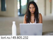 A dedicated and focused young woman with beautiful curly hair is diligently using a laptop in a stylish, modern workspace designed to enhance productivity and remote work options for everyone. A Young Woman Engaged Actively in Her Online Work Using a Laptop in a Modern Workspace Environment. Стоковое фото, агентство Ingram Publishing / Фотобанк Лори