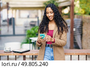 A cheerful young woman with curly hair smiles while texting on her smartphone at a cozy outdoor caf. A young woman is sitting outdoors, enjoying a warm cup of coffee while using her smartphone. Стоковое фото, агентство Ingram Publishing / Фотобанк Лори