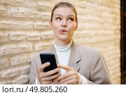 A youthful woman, wearing a contemplative and pensive look on her face, is absorbed in using her smartphone while casually leaning against a rugged and textured brick wall in broad daylight. A thoughtful woman is using her smartphone while standing against a textured brick wall outside. Стоковое фото, агентство Ingram Publishing / Фотобанк Лори