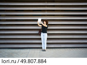 A joyful woman with curly hair holds her smartphone high, ready for a selfie in front of an elegant wall, capturing the vibrant essence of urban life and the pure joy from her radiant smile. A Cheerful Woman Taking a Fun Selfie Against a Modern Textured Wall with a Big Smile. Стоковое фото, агентство Ingram Publishing / Фотобанк Лори