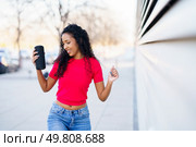 A joyful woman with curly hair dances exuberantly in an urban setting, holding a drink. Wearing a stylish red top and trendy jeans, she radiates fun and positivity, celebrating happiness. A Young Woman Joyfully Enjoys Music Outdoors in a Trendy Outfit, Reflecting Freedom and Fun. Стоковое фото, агентство Ingram Publishing / Фотобанк Лори