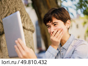 A young woman is having a delightful video call outdoors, where the beautiful natural setting creates a bright ambiance, as she covers her mouth with laughter and delight, expressing her joy. Joyful Interaction A Young Woman Laughing and Enjoying a Video Call Outdoors on a Beautiful Day. Стоковое фото, агентство Ingram Publishing / Фотобанк Лори
