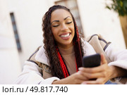 A cheerful young woman with curly hair and red braids beams joyfully as she uses her smartphone outdoors, capturing the essence of modern lifestyle and technology in todays digital age. A Young Woman is Enjoying Her Time on Her Smartphone While Relaxing Outdoors Under the Sun. Стоковое фото, агентство Ingram Publishing / Фотобанк Лори