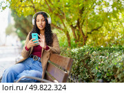 A young woman in trendy attire and stylish headphones sits on a park bench, engrossed in her smartphone, enjoying the peaceful atmosphere of the lush greenery and vibrant surroundings. A Young Woman Is Relaxing in the Park While Enjoying Music on Her Smartphone Device. Стоковое фото, агентство Ingram Publishing / Фотобанк Лори