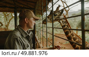 A man standing near a fence is reaching out to feed a giraffe on the other side. The giraffe is stretching its neck to eat from the mans hand. Стоковое AI-изображение, AI-художник Данила Большаков / Фотобанк Лори