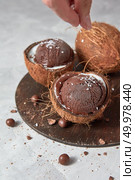 Two coconut halves with frozen fresh brown dessert and whole coconut with woman hand on a round wooden board on a gray stone table with copy space. Summer concept eating. Female hand coconut sprinkles on chocolate ice cream in coconut halves on a gray stone background. Стоковое фото, агентство Ingram Publishing / Фотобанк Лори