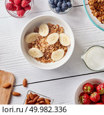 Grain free oat free paleo granola mixed with bananas slice, in bowl with nuts, raspberries, strawberries, blueberries and milk in a decanter on a white wooden background. Healthy breakfast ingredients on a white woodwn background. Granola, nuts, berries, milk, banana in bowl , top view. Стоковое фото, агентство Ingram Publishing / Фотобанк Лори