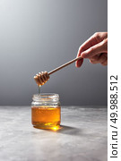 Woman hand hold a wooden stick with dripping flower honey into a glass jar on a gray background, place for text. Pure organic sweet goodness.. Wooden stick with natural flower honey in a womans hand above a gray concrete table. Rosh hashanah jewish New Year holiday. Стоковое фото, агентство Ingram Publishing / Фотобанк Лори