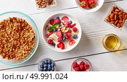 A ready breakfast of natural organic ingredients - granola, berries, muesli, nuts, honey, milk for the preparation of natural healthy food. Concept of organic eating.. Cereal muesli and sliced bananas, strawberries, blueberries, raspberries, almonds and walnuts with milk in white ceramic bowl on white table. Top view. Стоковое фото, агентство Ingram Publishing / Фотобанк Лори