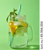 Summer natural sparkling fresh lemonade in mason glass jar with large bubbles of air on a green background. Concept of cold alcoholic or non-alcoholic summer drinks.. Refreshing carbonated homemade drink with natural ingredients - citrus fruits, ice cubes, green mint, colored plastic straw in a glass jar on a green background. Стоковое фото, агентство Ingram Publishing / Фотобанк Лори