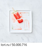 Frozen juice on a stick. Broken ice cream in a plate with splashes of ice cream on a gray marble background with copy space. Flat lay. Broken fruit ice cream lolly on a white plate with a pattern from the thawing ice cream on a gray marble background. Copy space for text. Flat lay. Стоковое фото, агентство Ingram Publishing / Фотобанк Лори
