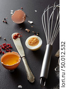 Knife with red pepper, a metal whisk and halves of raw and boiled eggs on a black concrete background. Set for cooking. Raw and boiled egg, a metal whisk and a knife with red pepper on a black concrete background. Preparation. Стоковое фото, агентство Ingram Publishing / Фотобанк Лори