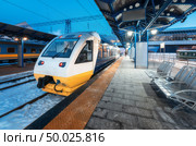 High speed train on the railway station at night in Europe. Urban landscape with modern commuter train on the railway platform with illumination at dusk. Intercity vehicle. Passenger railroad travel. Стоковое фото, агентство Ingram Publishing / Фотобанк Лори