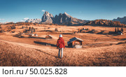 Young woman on the hill looking on meadows and mountains at sunset in autumn. Alpe di Siusi, Dolomites, Italy. Landscape with girl in red jacket, field with yellow grass, wooden houses and rocks. Стоковое фото, агентство Ingram Publishing / Фотобанк Лори